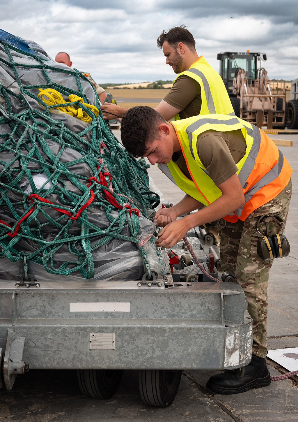 Personnel from 1 Air Mobility Wing, in camouflage trousers and a high-visibility vest secure a large cargo pallet wrapped in green netting on an airfield. The pallet is destined to be loaded onto a Atlas (A400M) aircraft.
